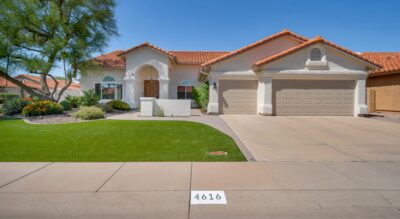 A well-maintained suburban home with a beige stucco exterior, a tile roof, a neatly manicured green lawn, desert landscaping, and a three-car garage at 4616 E Gelding Dr.